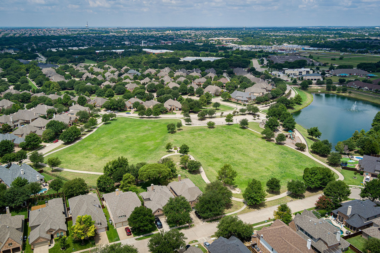 Aerial view of a suburban neighborhood featuring rows of houses, a large central green park, walking paths, and a nearby pond under a partly cloudy sky.