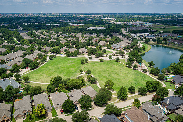 Aerial view of a suburban neighborhood with rows of houses, a large green park in the center, walking paths, trees, and a pond on the right.