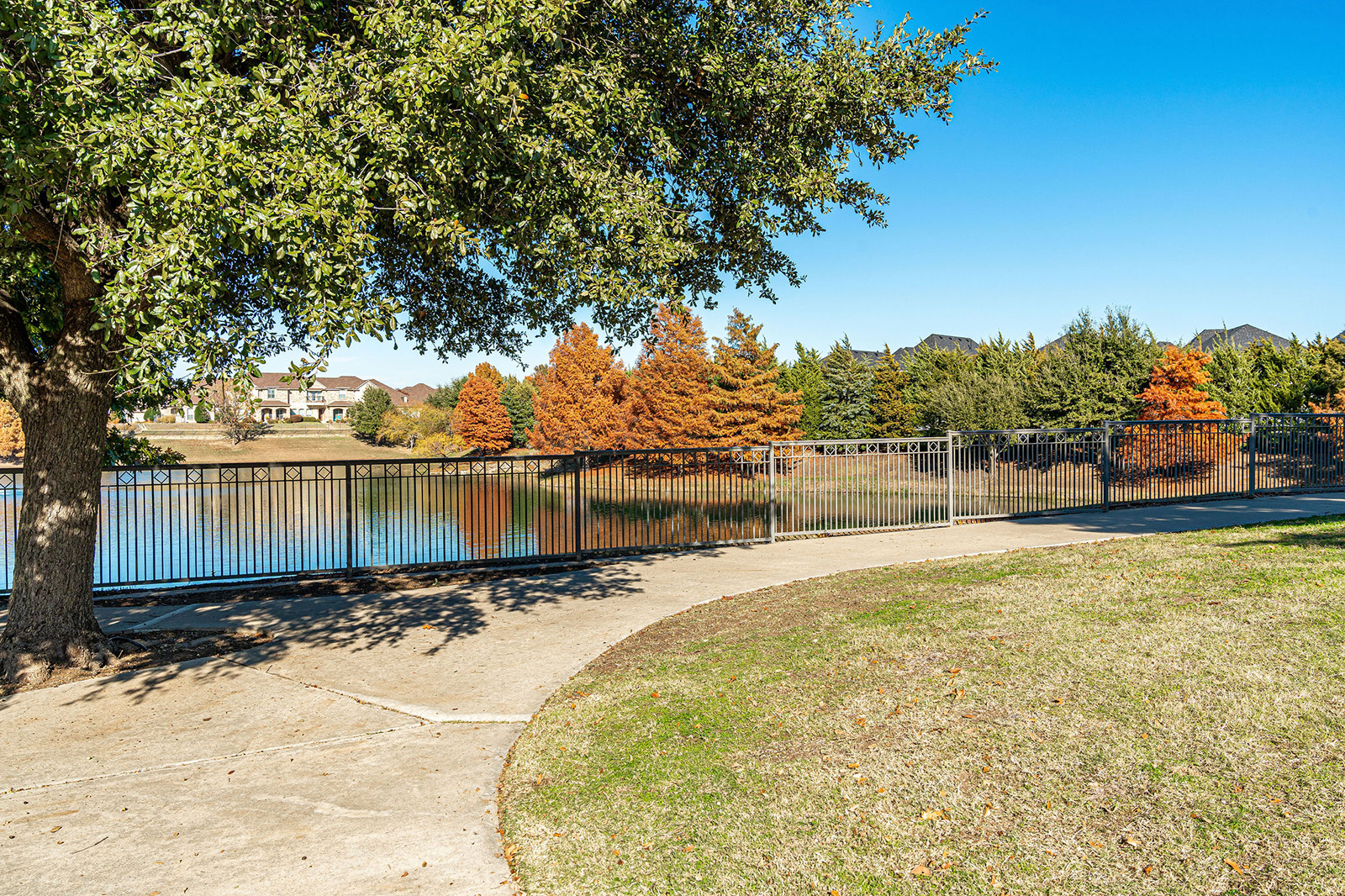 A paved pathway curves beside a metal fence overlooking a calm lake, with trees and houses visible in the background on a clear day.