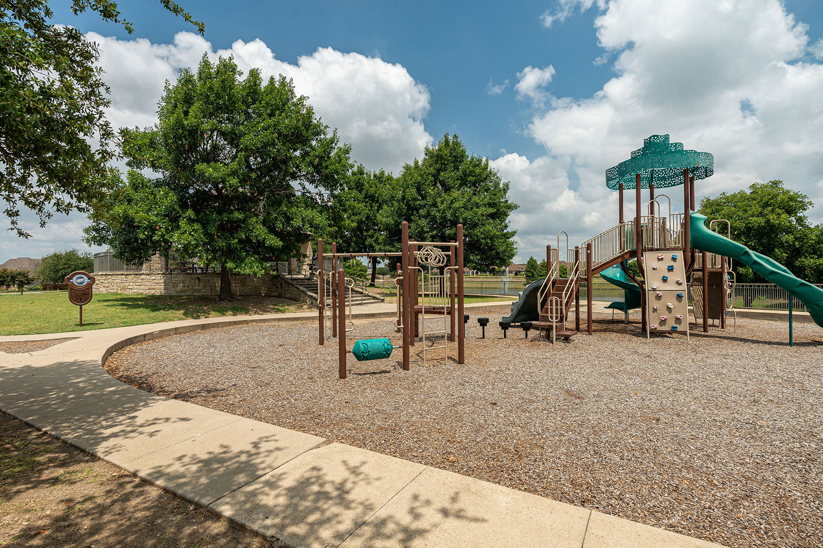Outdoor playground with climbing structures, slides, and a small rock wall, surrounded by trees and a walking path on a partly cloudy day.