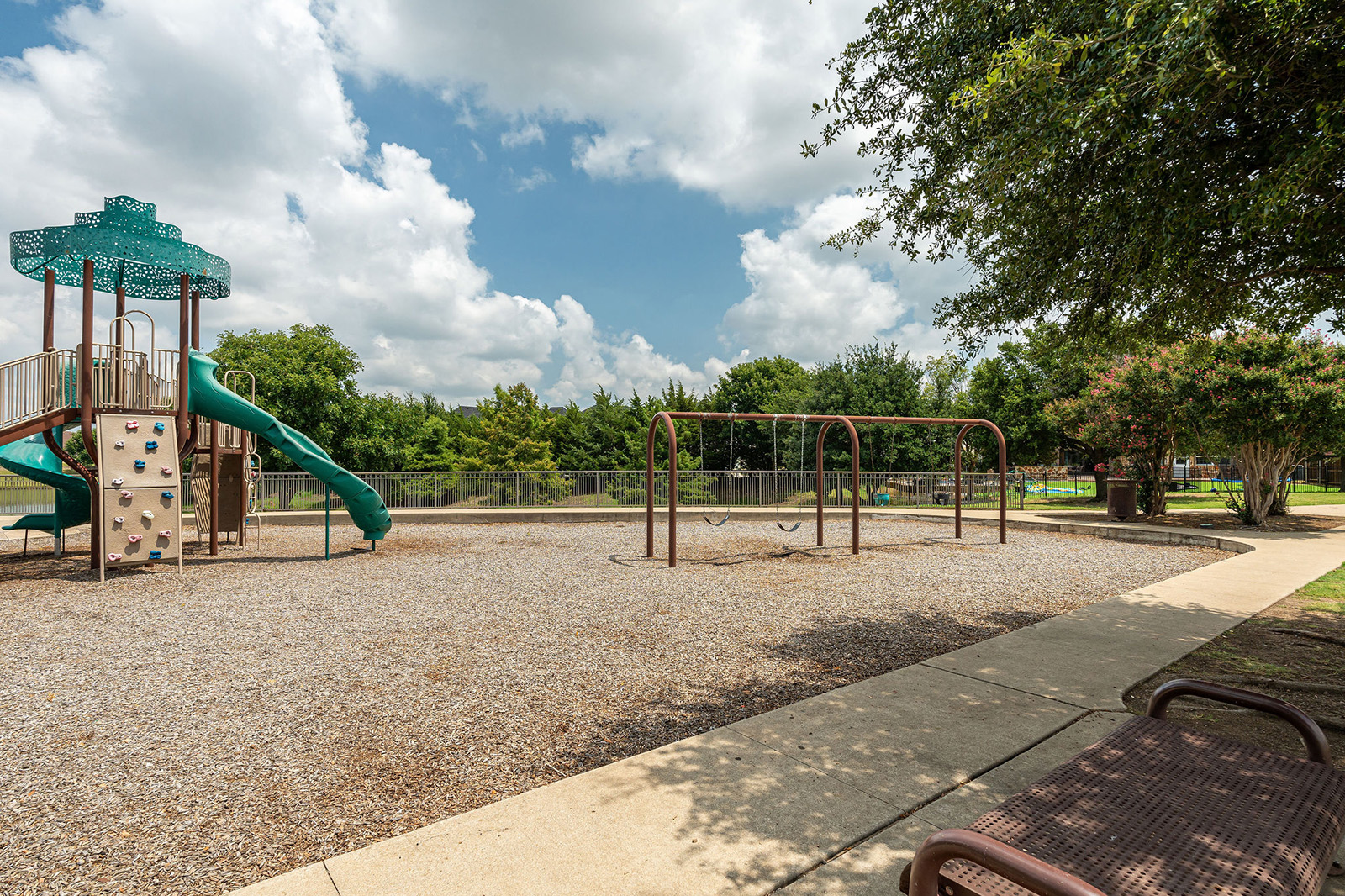 A playground with a green slide, climbing wall, swing set, and gravel ground, bordered by a sidewalk and trees under a partly cloudy sky.