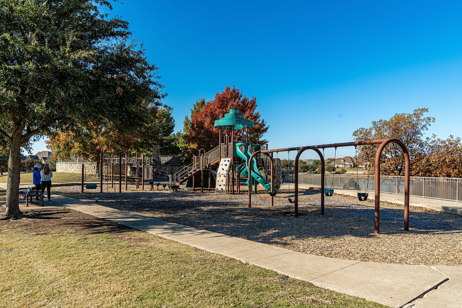 A playground with swings, slides, and climbing structures, surrounded by trees and a fence. Two people stand near a tree on the left. The sky is clear and blue.