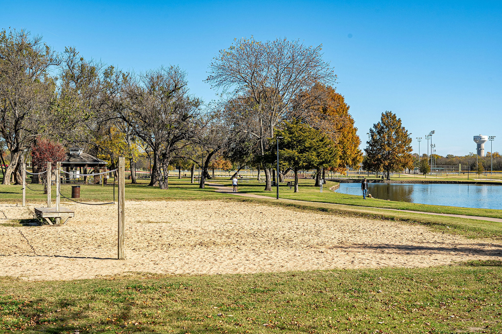 A sandy volleyball court in a park with trees, grassy areas, a walking path, and a pond; people are walking and enjoying the outdoor setting on a clear day.