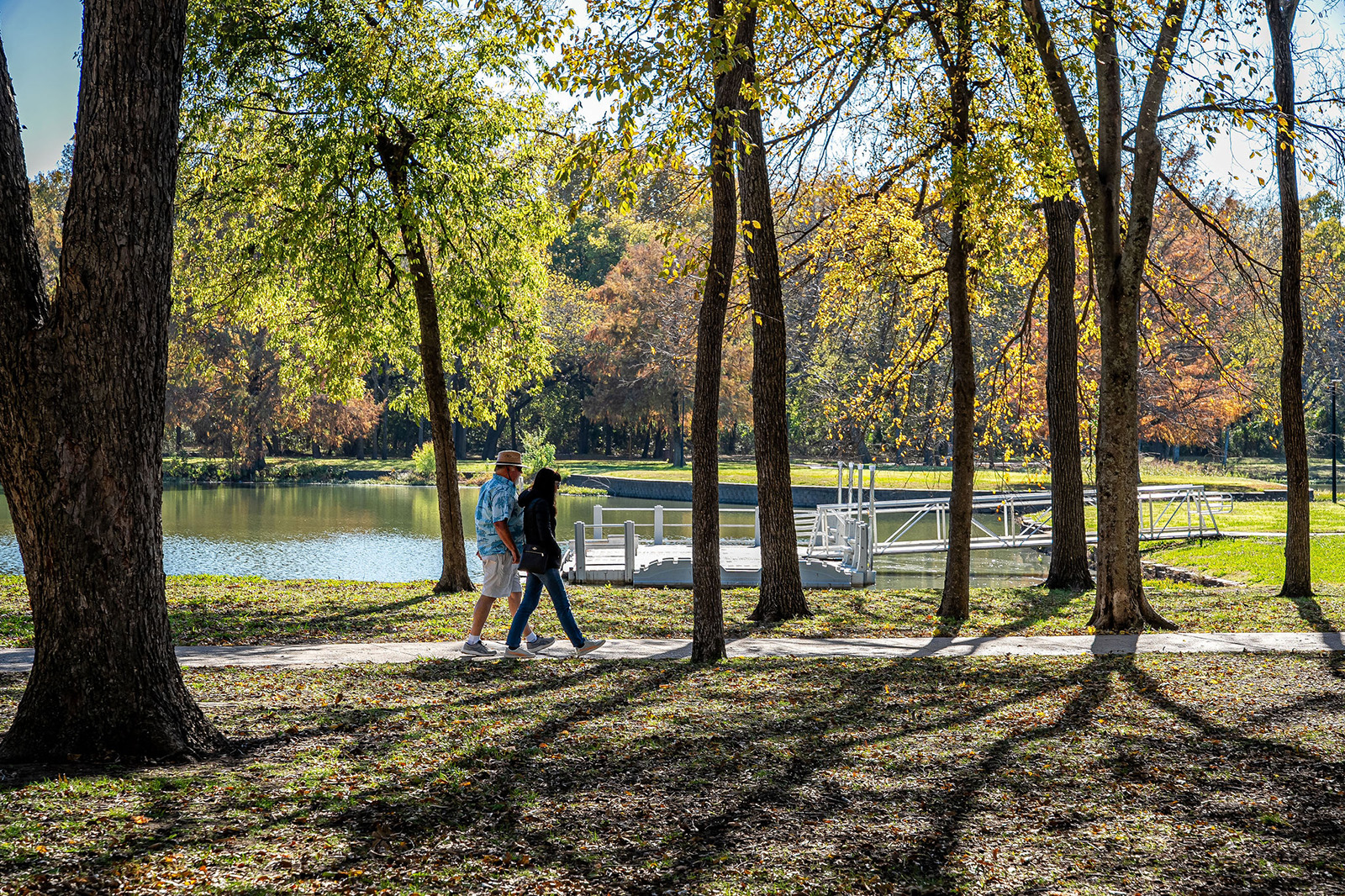 Two people walk on a shaded path beside a lake with a dock, surrounded by trees with sunlight filtering through the branches.