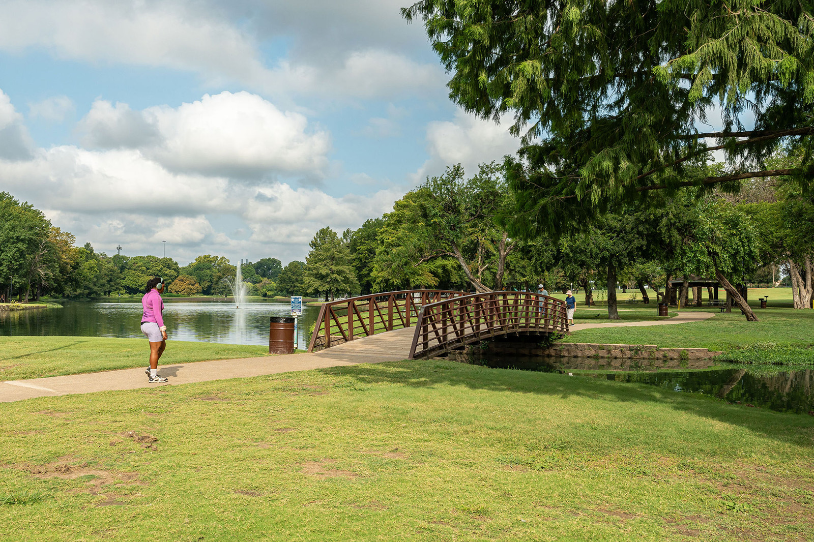 A person walks on a paved path near a small arched footbridge over a creek in a green park with a pond and fountain in the background.