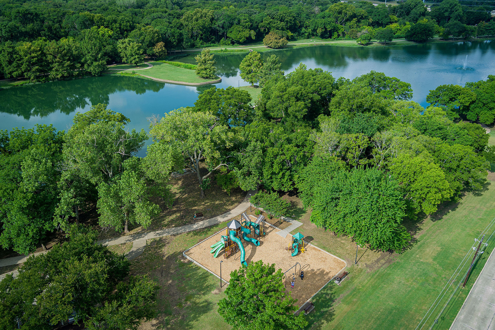 Aerial view of a playground surrounded by trees near a lake, with walking paths and grassy areas visible.
