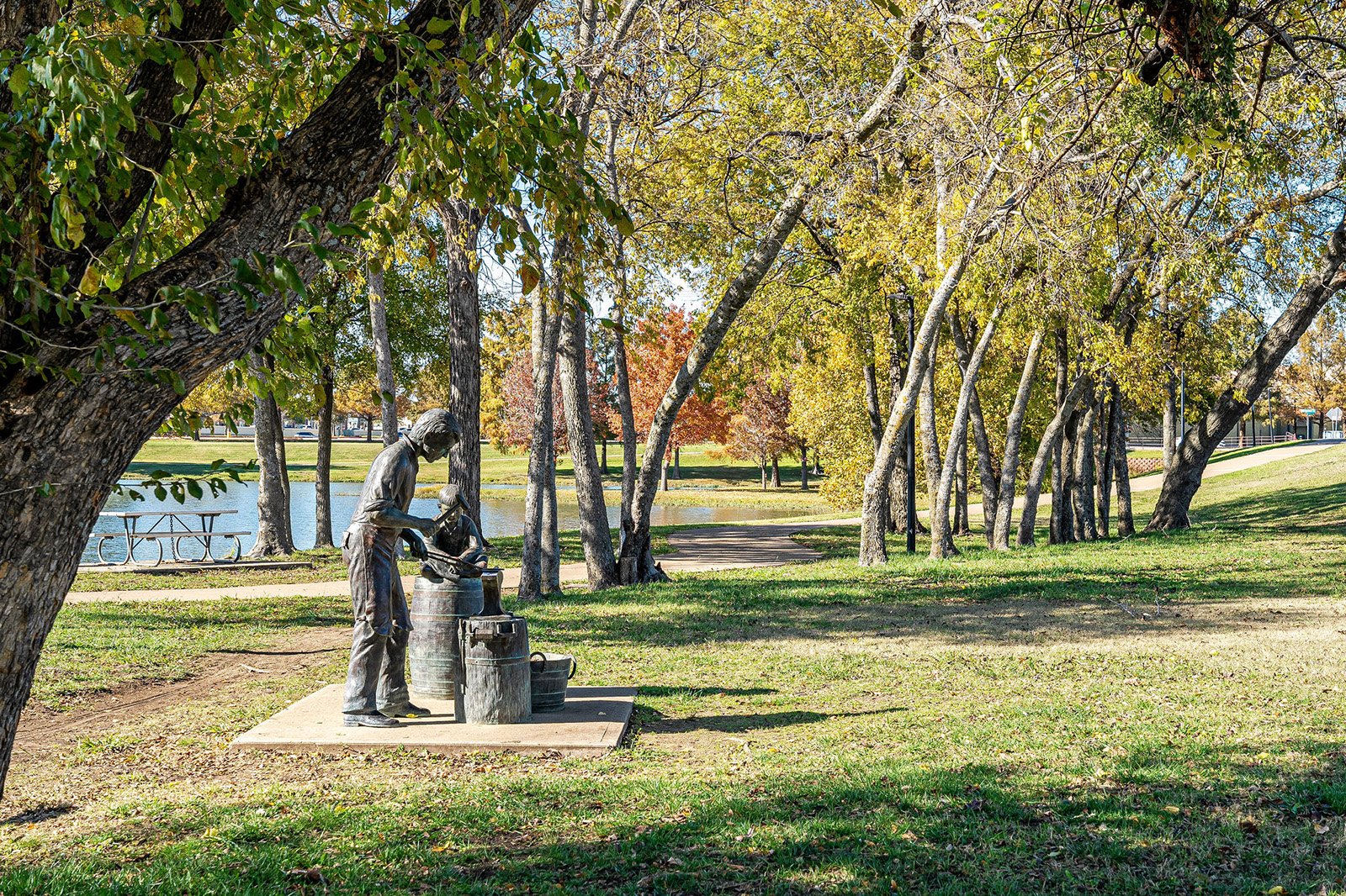 A bronze statue of a person stands beside barrels in a tree-lined park near a pond, with sunlight filtering through autumn foliage.