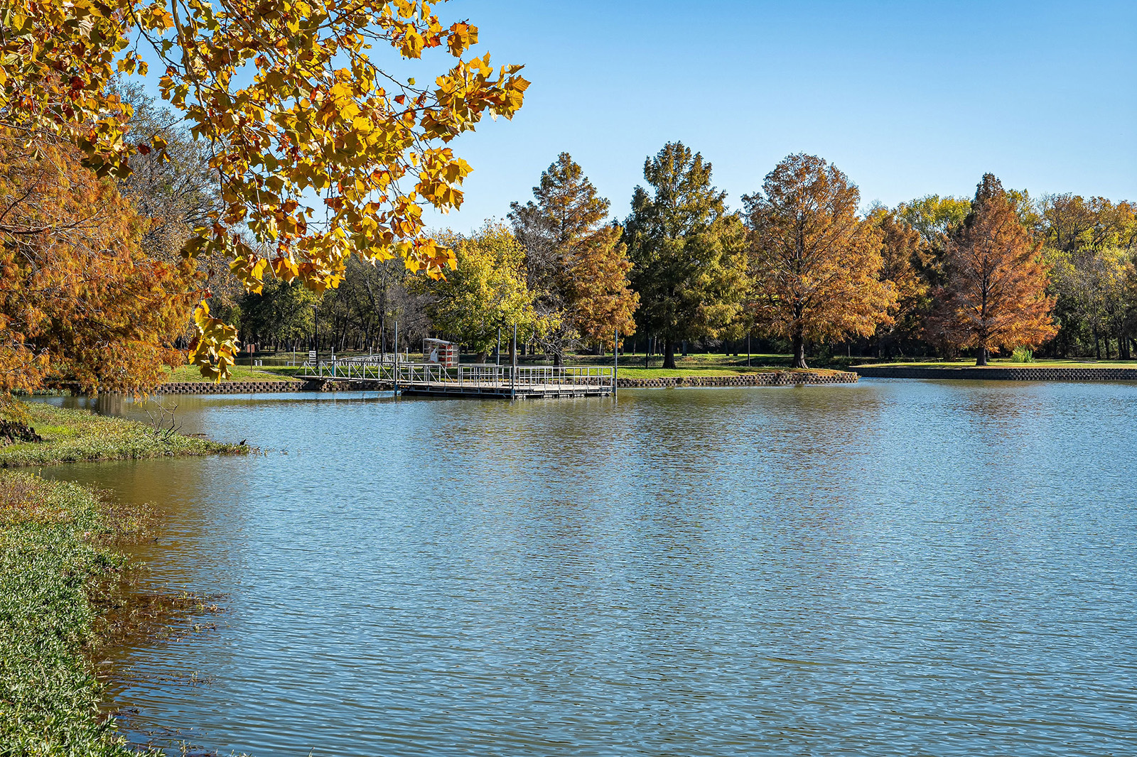 A calm lake with a dock, surrounded by trees with autumn foliage under a clear blue sky.