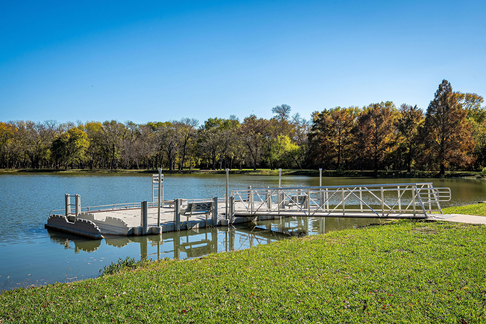 A floating dock with a ramp extends into a calm lake, surrounded by green grass and trees under a clear blue sky.