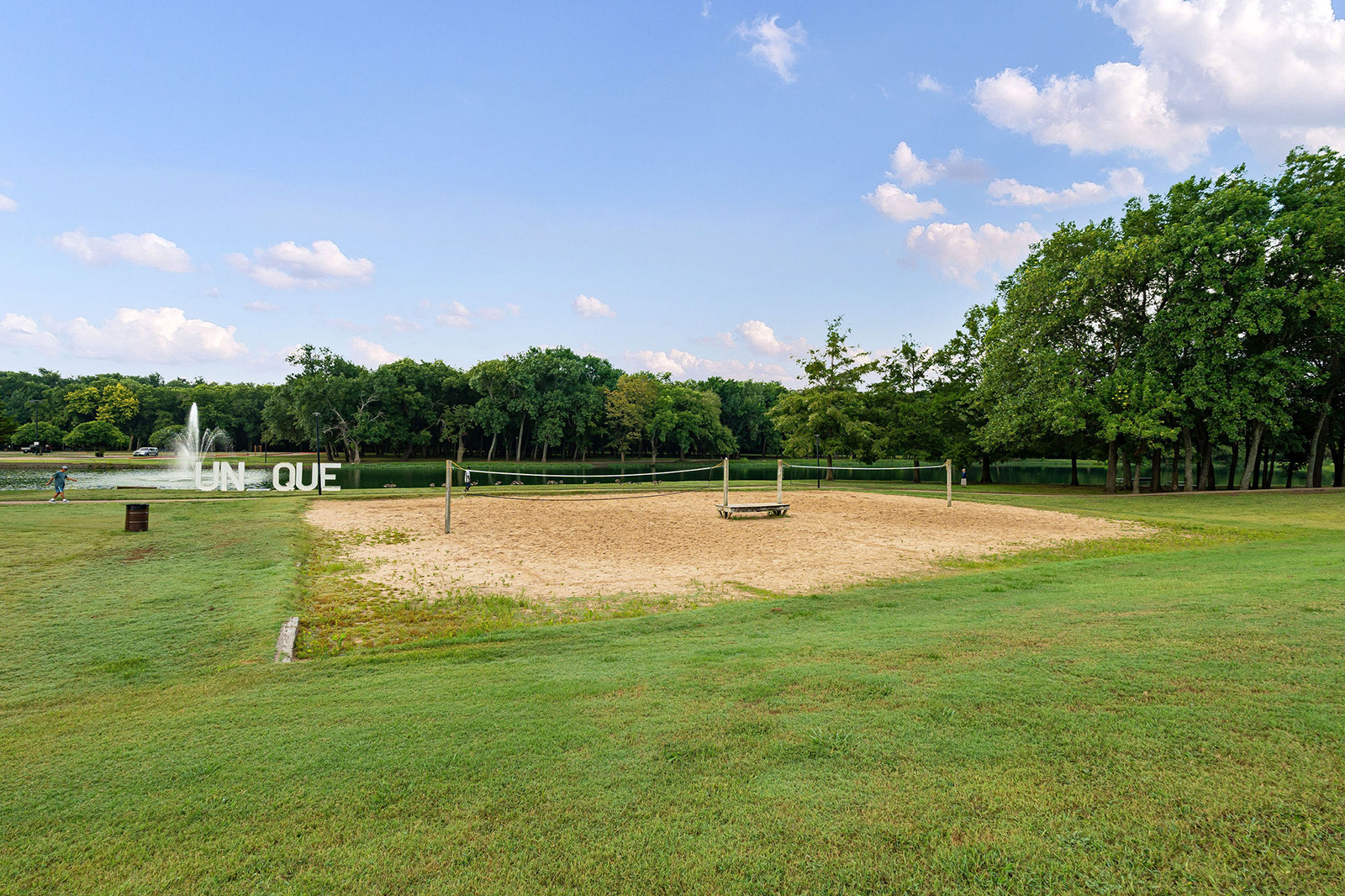 A sand volleyball court sits on a grassy field near trees, with a pond and fountain in the background on a partly cloudy day.