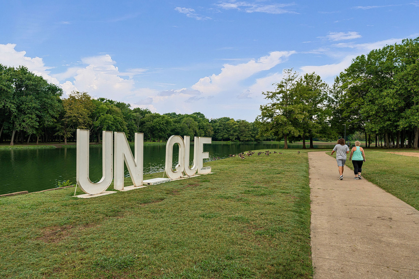 Two people walk on a paved path by a lake, passing large white letters spelling “UN QUE” on the grassy shore; trees line the water and sky is partly cloudy.