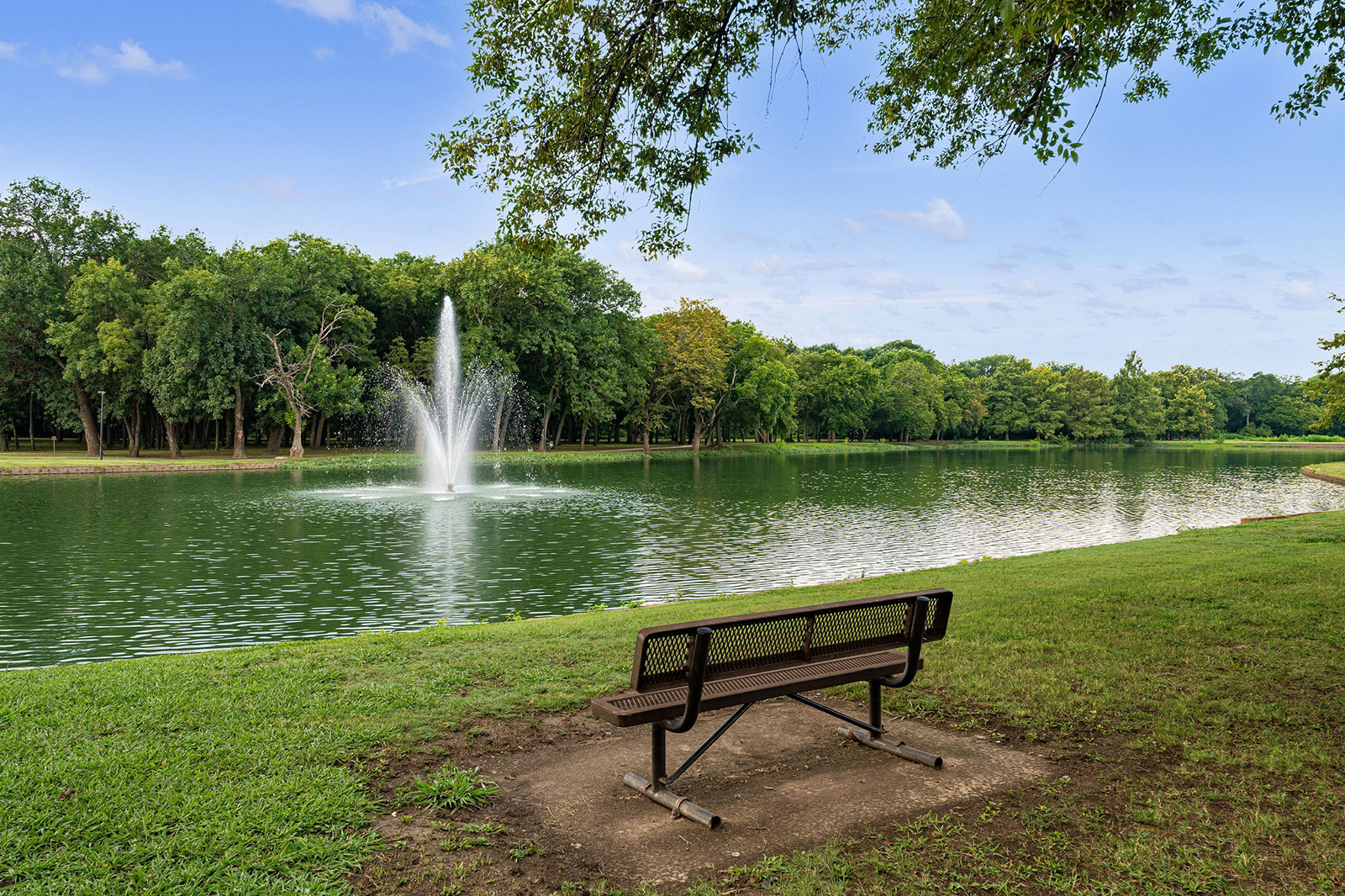 A metal bench faces a pond with a water fountain in the center, surrounded by green grass and trees under a clear blue sky.