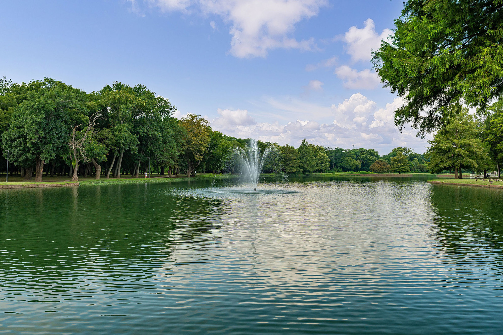 A calm pond with a water fountain in the center, surrounded by green trees under a partly cloudy sky.