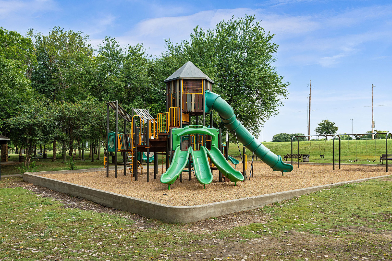 A playground structure with multiple green slides, climbing elements, and a covered tower, surrounded by trees and set on a mulch surface.