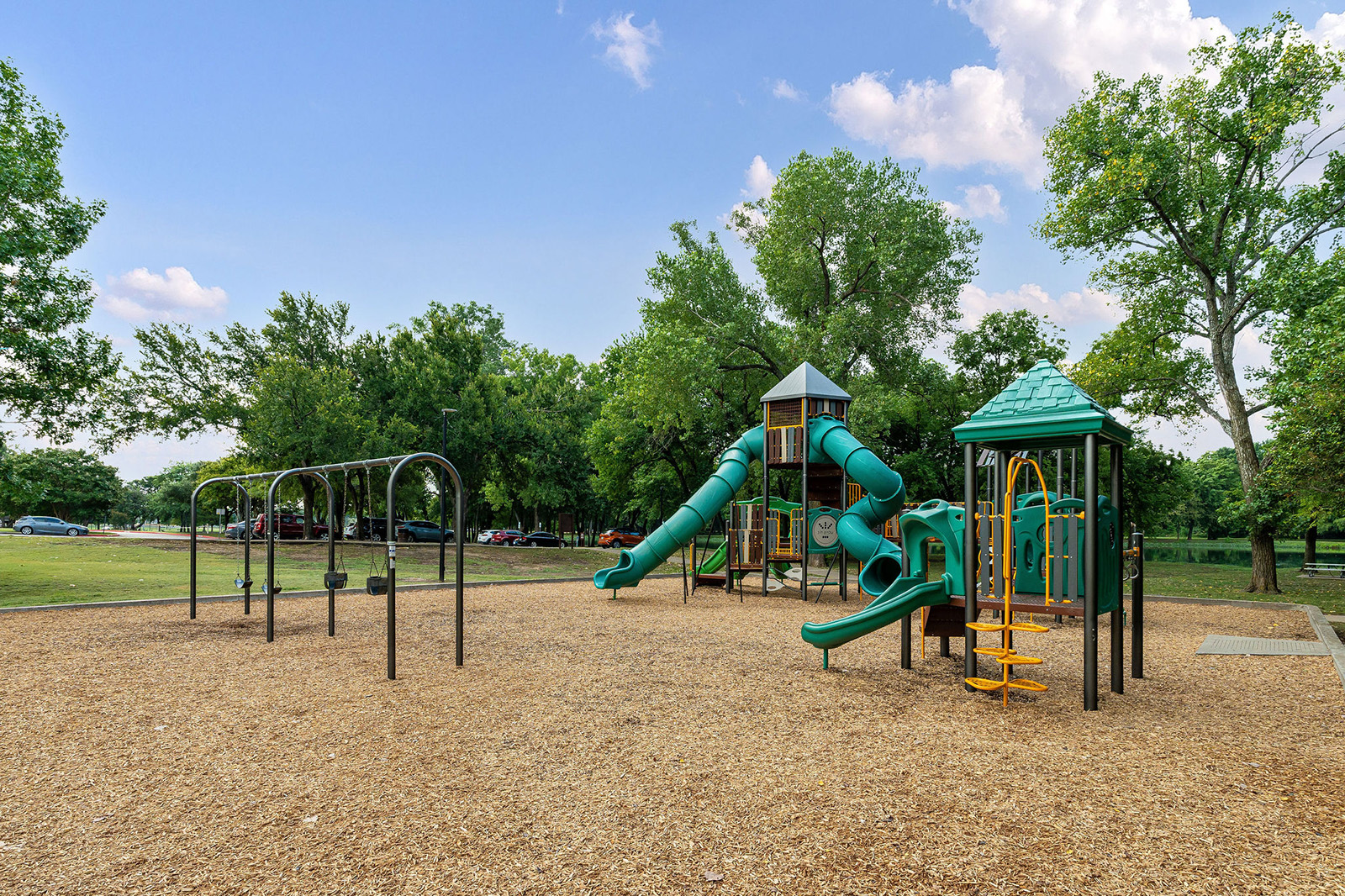 Outdoor playground featuring swings, slides, and climbing structures on a mulch surface, surrounded by green trees and an open grassy area under a blue sky.