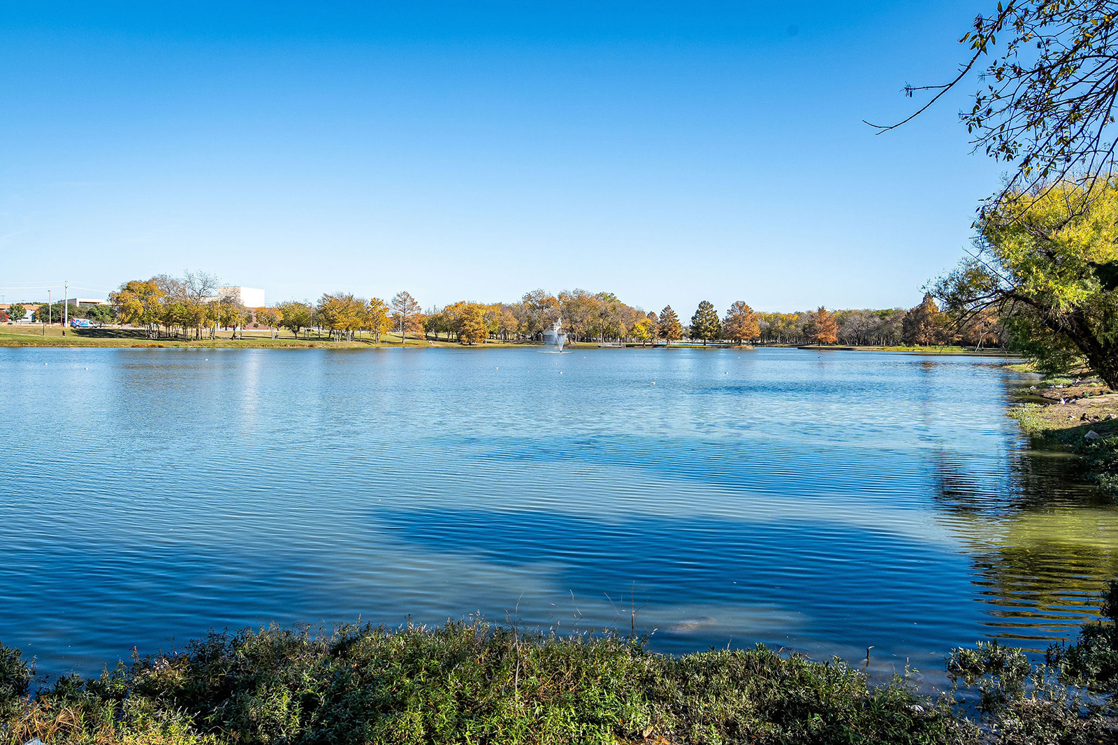 A clear lake with gentle ripples, bordered by trees with autumn foliage under a bright blue sky; a fountain is visible in the distance.