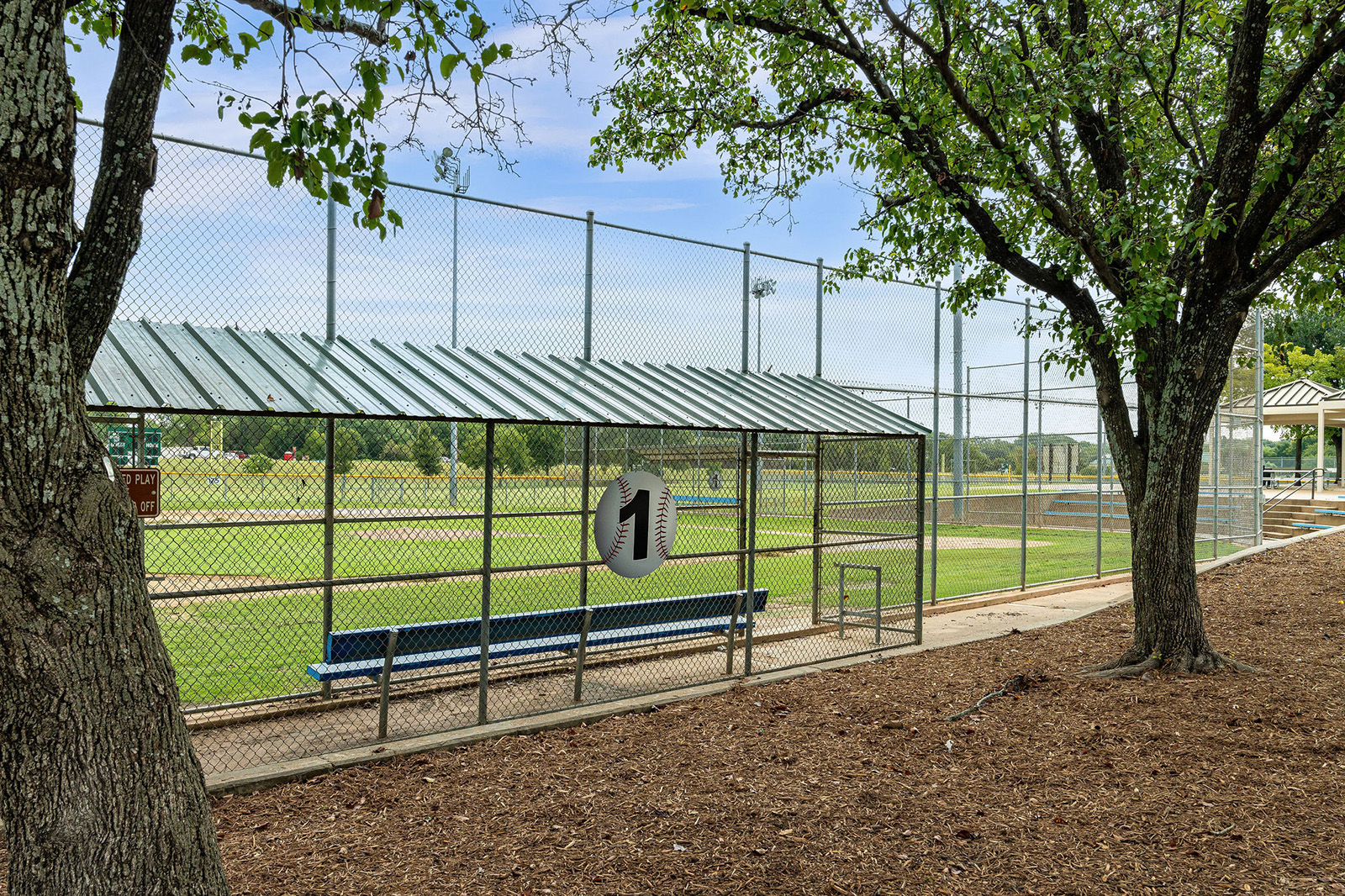 A fenced baseball dugout with a blue bench and metal roof sits near a field, surrounded by trees and open grassy areas.