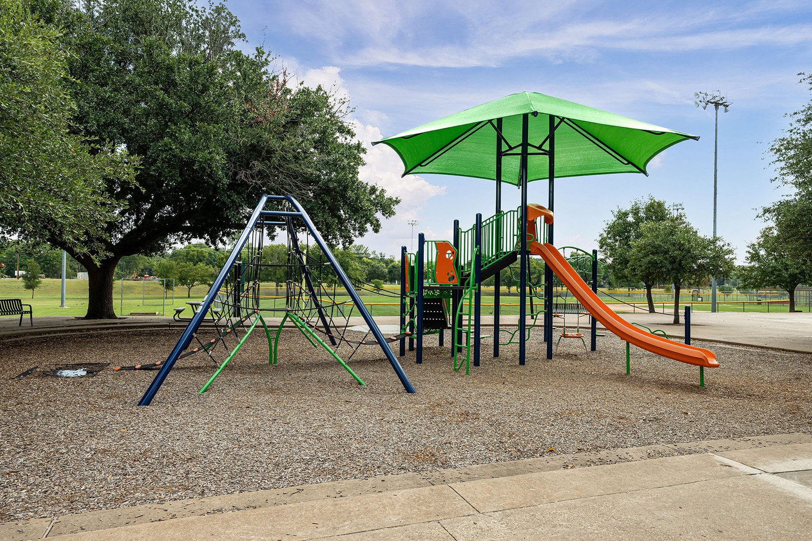 Playground with orange slides, a climbing structure, swings, and a large green canopy on a gravel surface, surrounded by trees and open grassy areas.