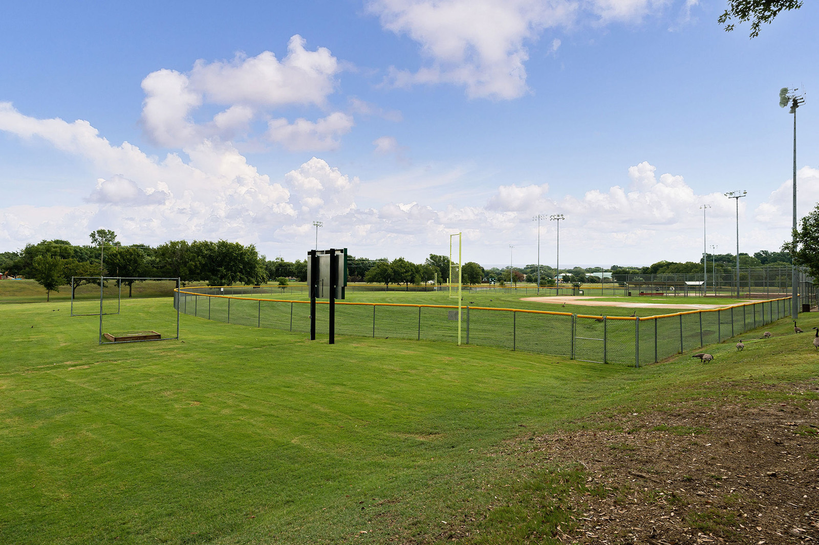 A fenced baseball field with yellow foul poles and green grass under a partly cloudy sky, surrounded by open parkland and tall light poles.