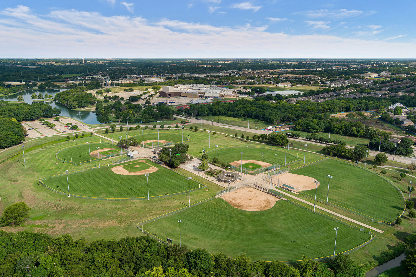 Aerial view of a sports complex with several baseball and softball fields, surrounded by green grass, trees, and nearby roads under a blue sky.