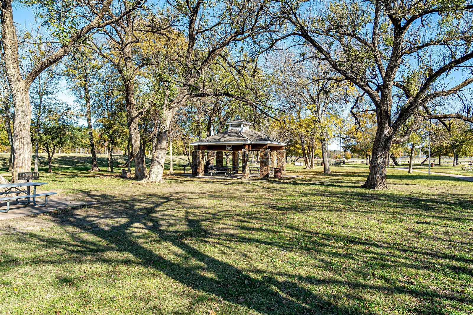 A stone gazebo stands among trees in a grassy park on a sunny day, with a picnic table nearby and long shadows cast on the lawn.