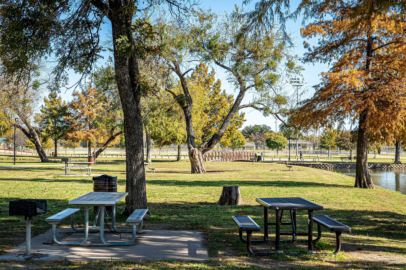 Picnic tables and benches under trees in a park with a grill, open grassy areas, a pond, and a wooden footbridge in the background.