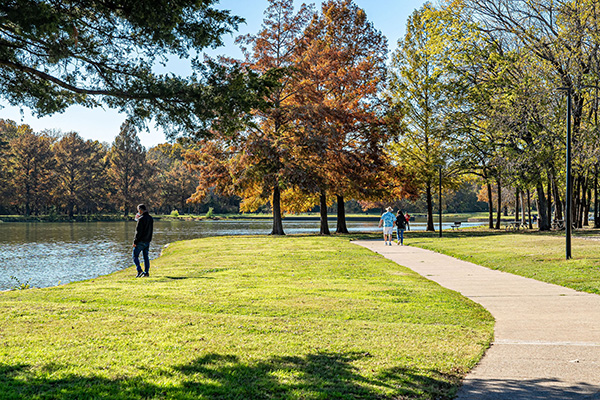 A paved path runs beside a lake with trees showing fall colors; a person walks alone while two others walk together in the distance.