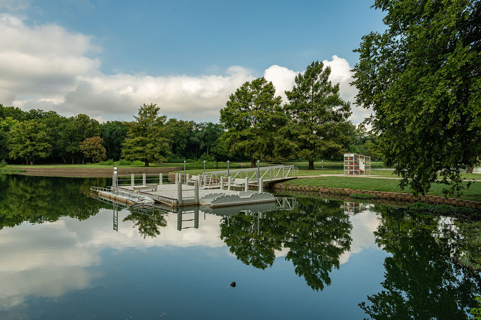 A floating dock extends over a calm lake surrounded by trees, with reflections visible in the water under a partly cloudy sky.