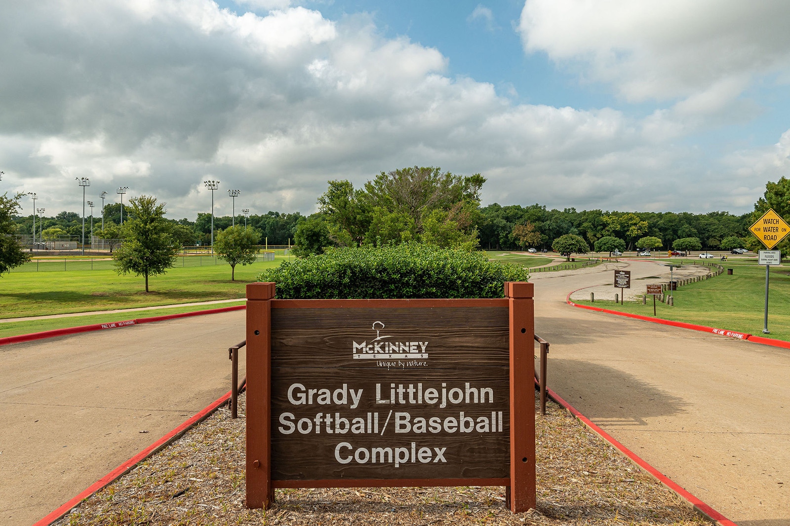 A wooden sign for Grady Littlejohn Softball/Baseball Complex stands at the entrance to a sports facility with fields and trees visible in the background.