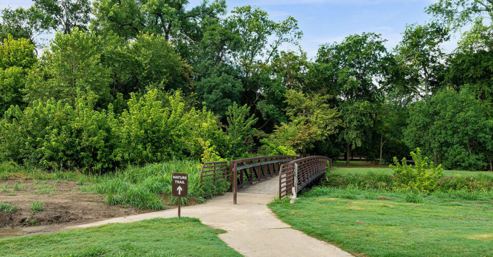 A paved path leads to a small metal footbridge surrounded by green trees, with a sign reading “Nature Trail” at the entrance.