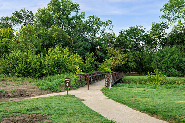 A paved sidewalk leads to a small metal bridge surrounded by green trees and grass, with a sign marking the walking trail.