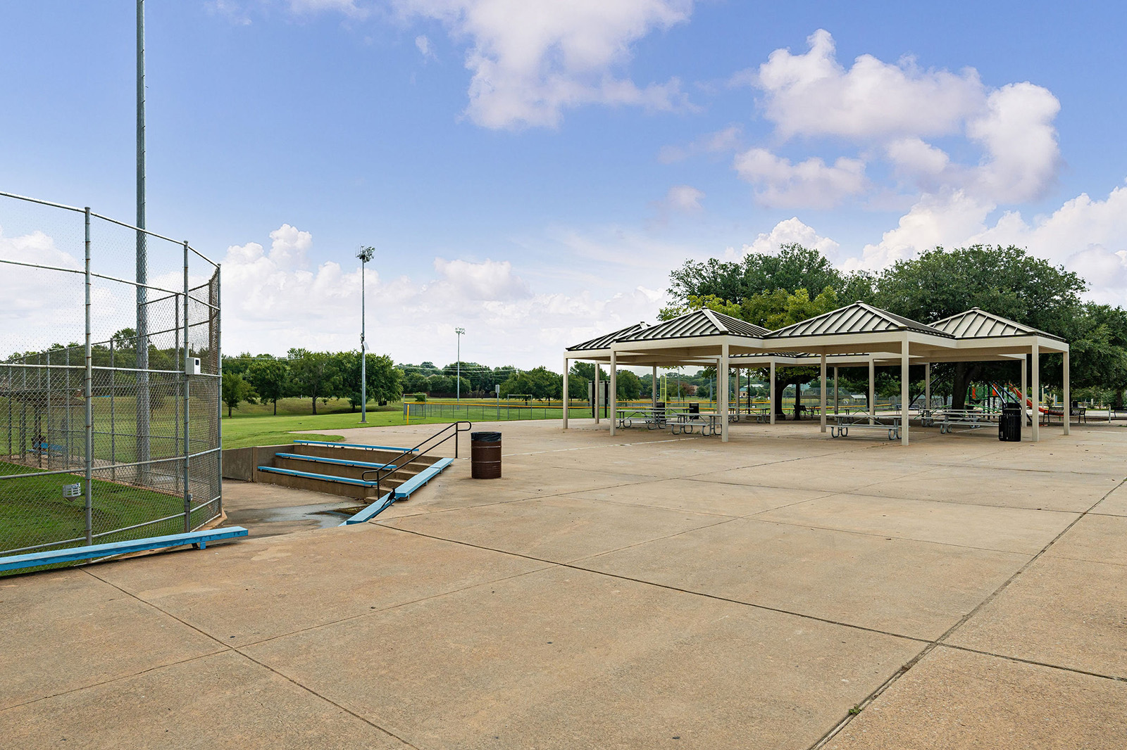 Outdoor park with a baseball field on the left, concrete seating steps, picnic pavilions with tables, and open grassy areas under a partly cloudy sky.