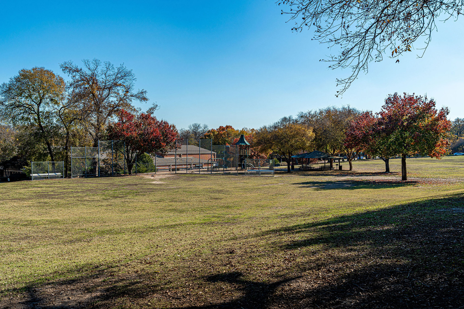 A grassy field with a playground, baseball backstop, and trees with autumn foliage under a clear blue sky.