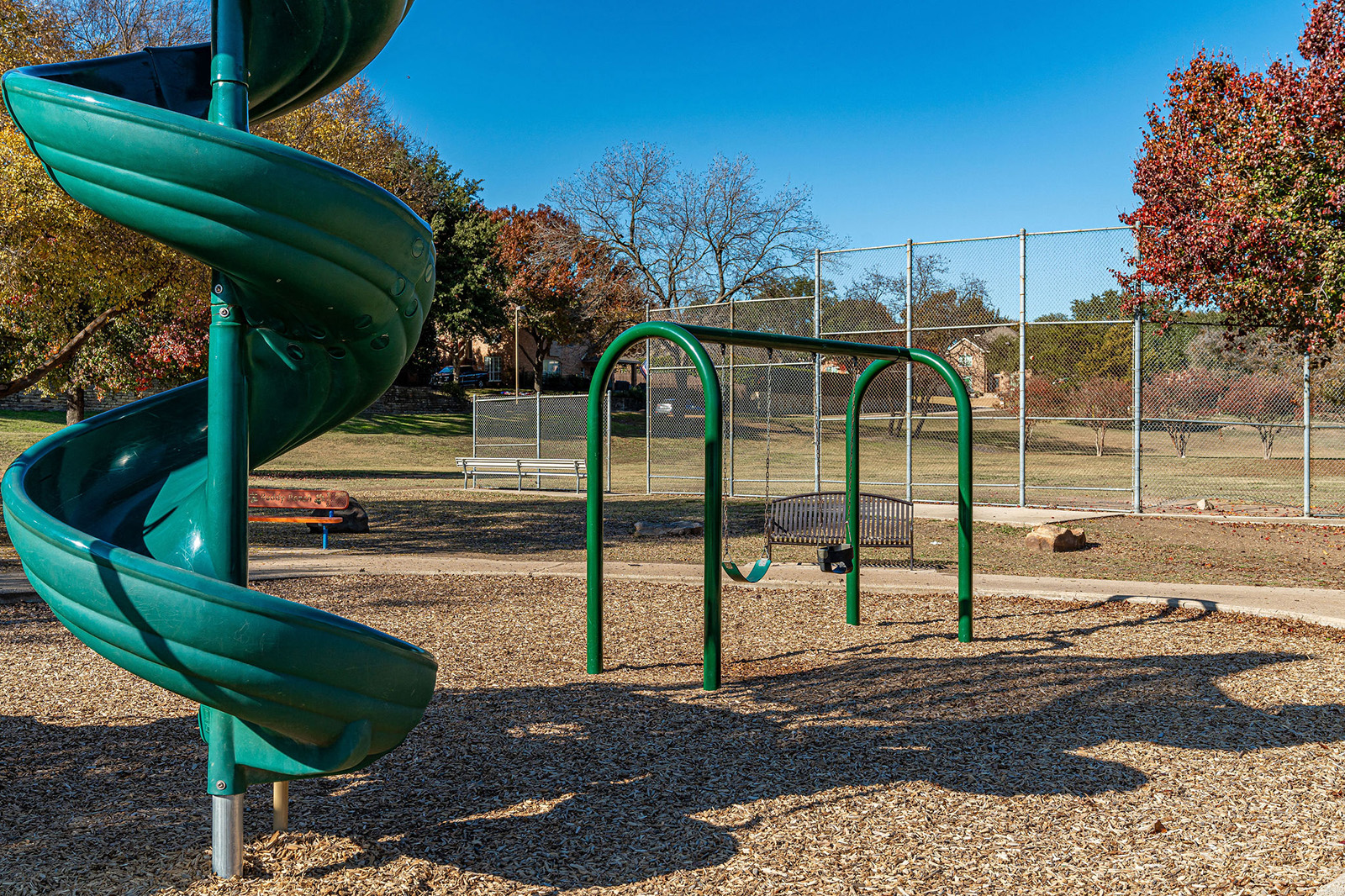 A playground with a green spiral slide, a swing set, benches, a baseball backstop, and trees, under a clear blue sky.