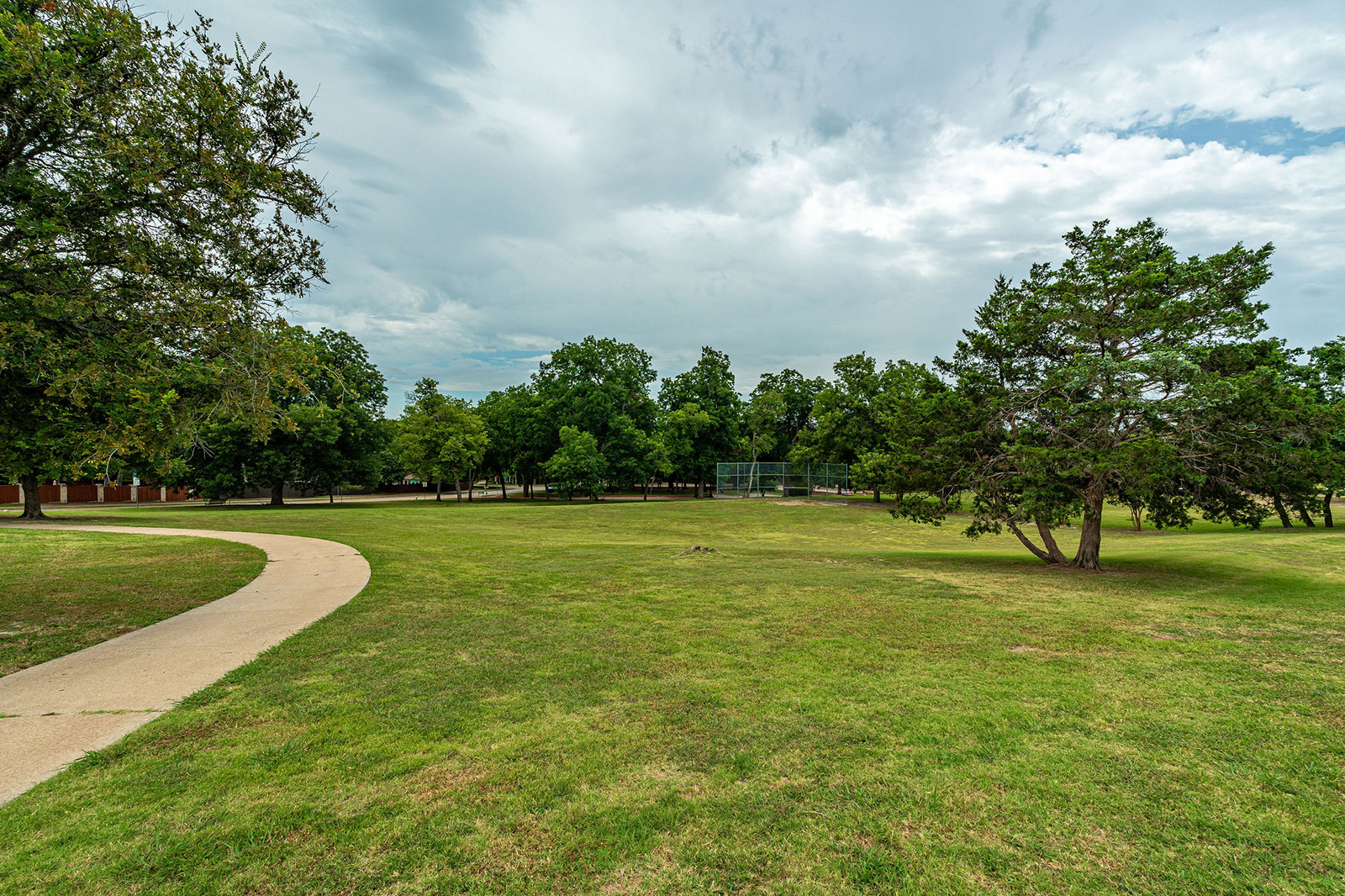 A paved path curves through a grassy park with scattered trees, under a cloudy sky. A fenced baseball backstop is visible in the distance.