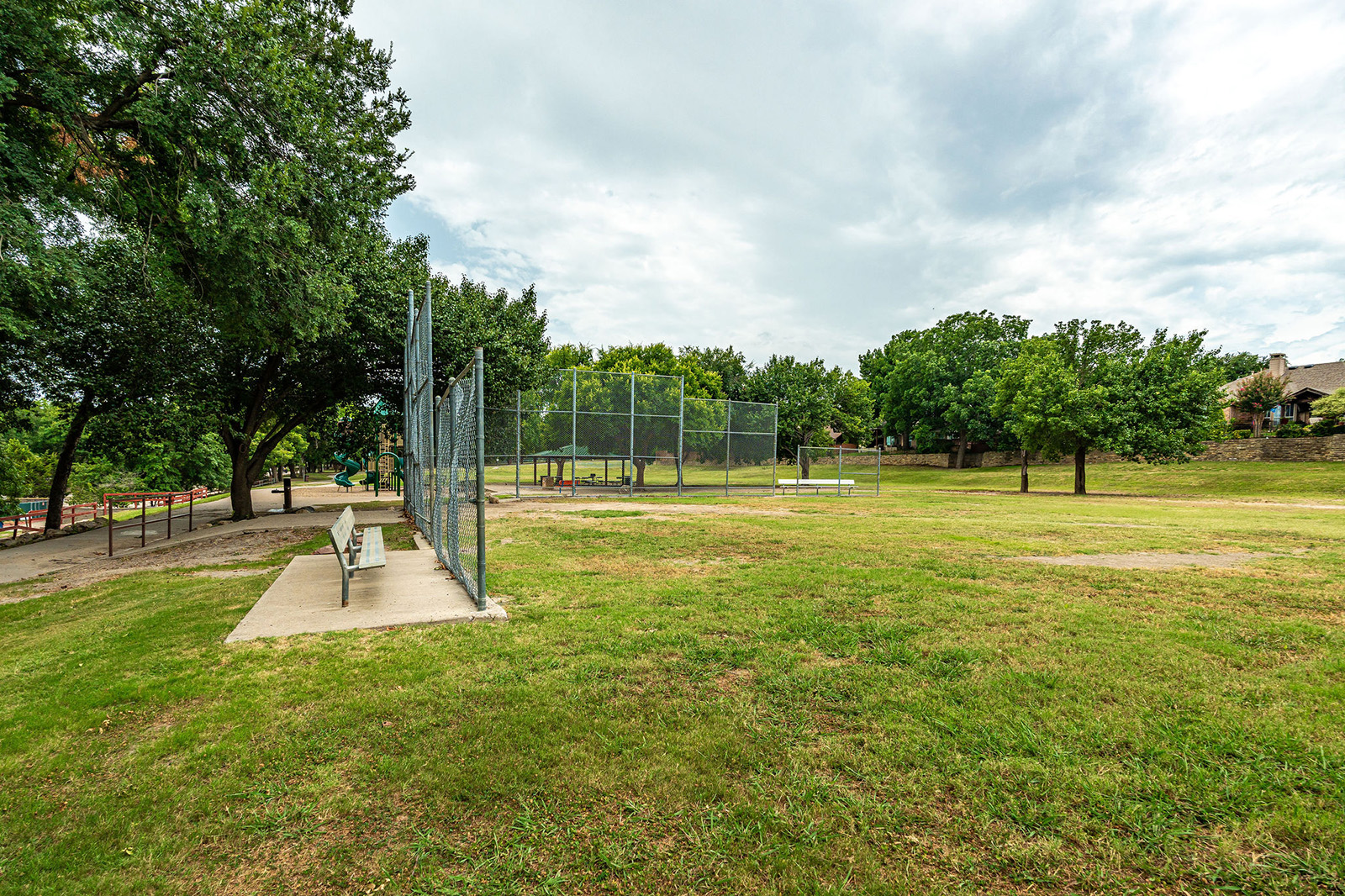 A small baseball field with a dugout and chain-link fence is surrounded by grass and trees in a suburban park.