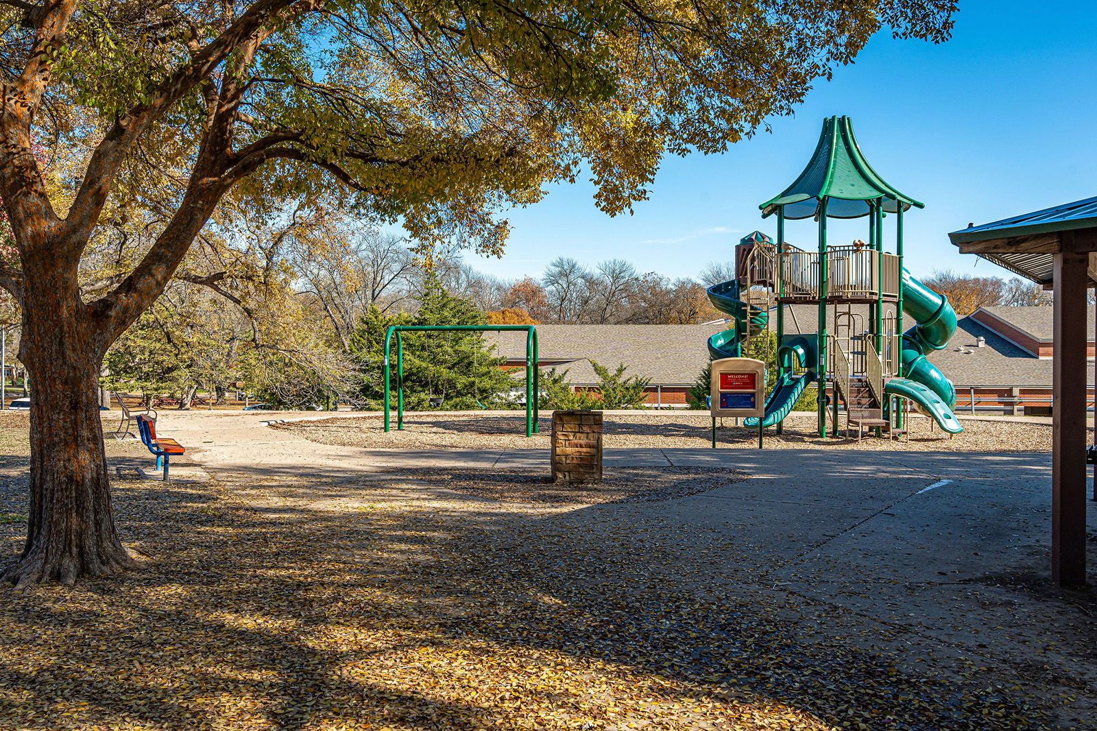Playground with green slides, swings, and climbing structures surrounded by trees with fallen leaves; residential houses are visible in the background.