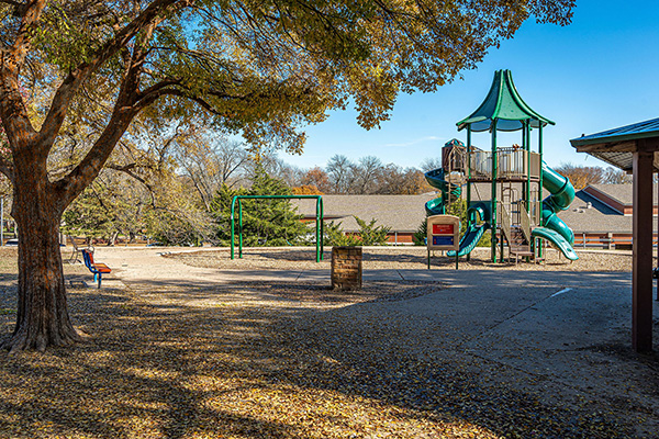 A playground with a tall green slide structure, swings, benches, and large trees casting shade on the ground covered in fallen leaves.