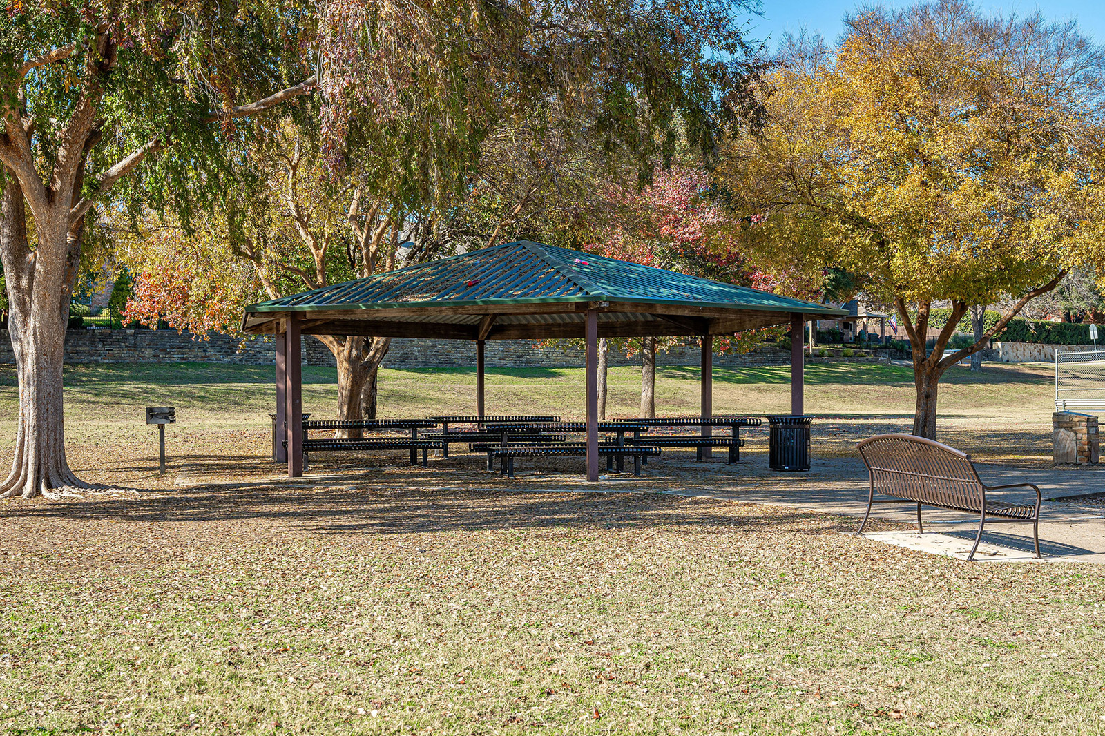 A covered picnic pavilion with tables and benches stands in a grassy park area surrounded by trees, a grill, and a nearby metal bench.