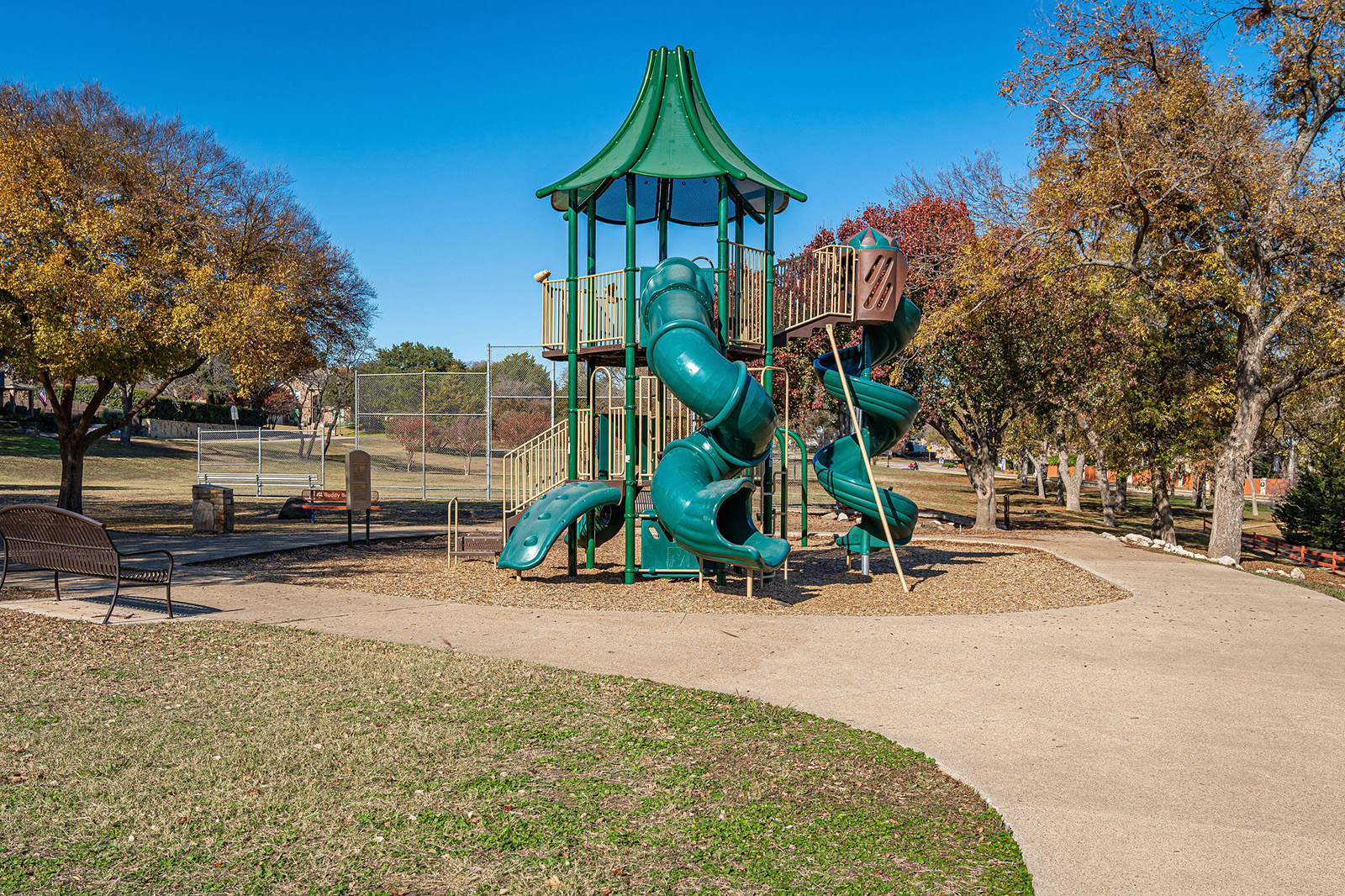 A green playground structure with spiral slides stands in a park surrounded by trees, benches, and a walking path under a clear blue sky.
