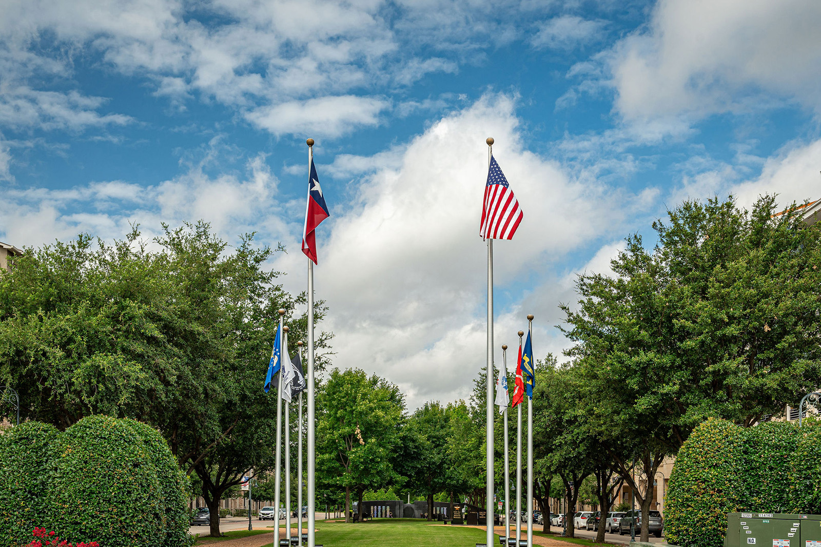Six flagpoles with various flags, including the Texas and U.S. flags, stand in a landscaped area bordered by trees under a partly cloudy sky.