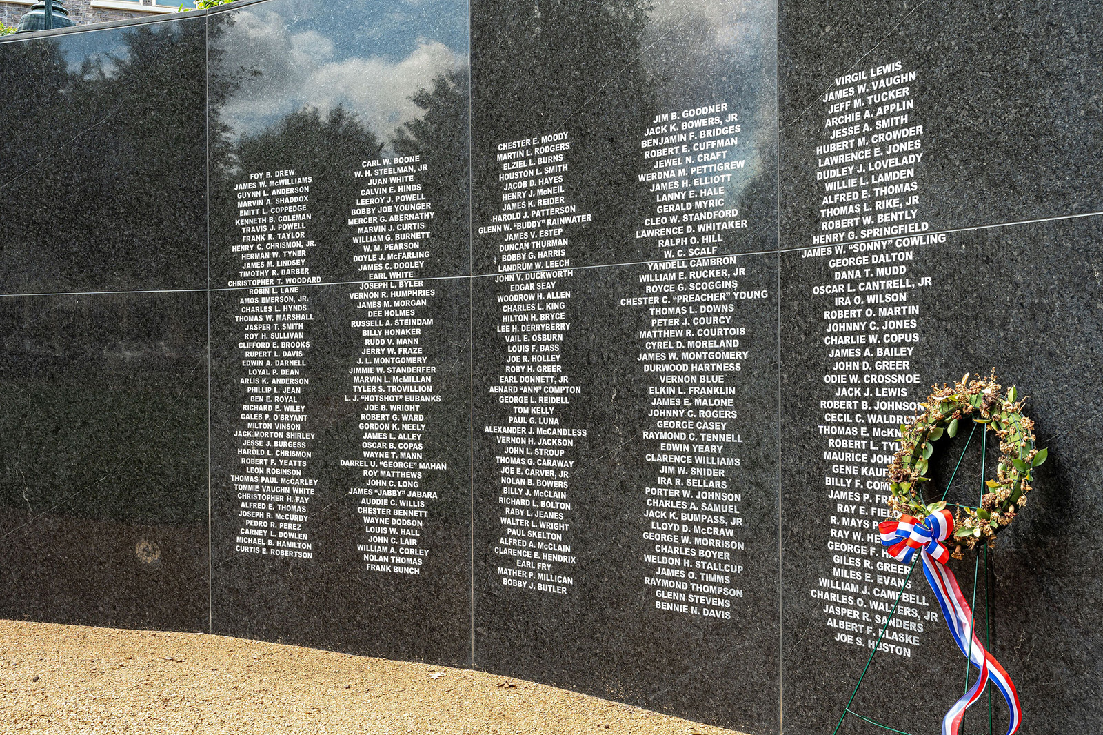 A granite memorial wall engraved with names, next to a gravel path. A wreath with red, white, and blue ribbon is placed at the base of the wall.