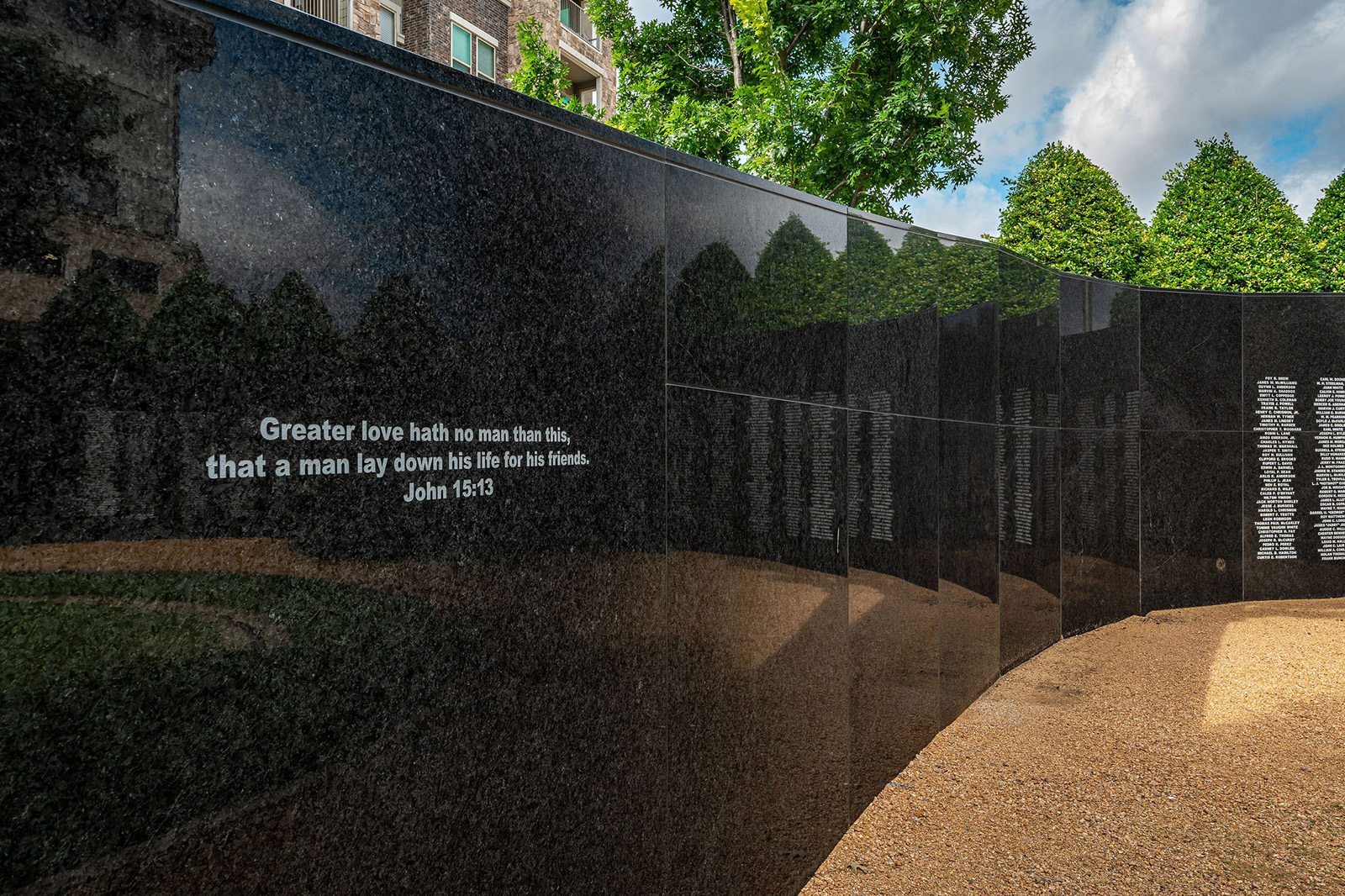 Black stone memorial wall engraved with John 15:13, reflecting trees and sky, with additional names listed further along the curved wall.