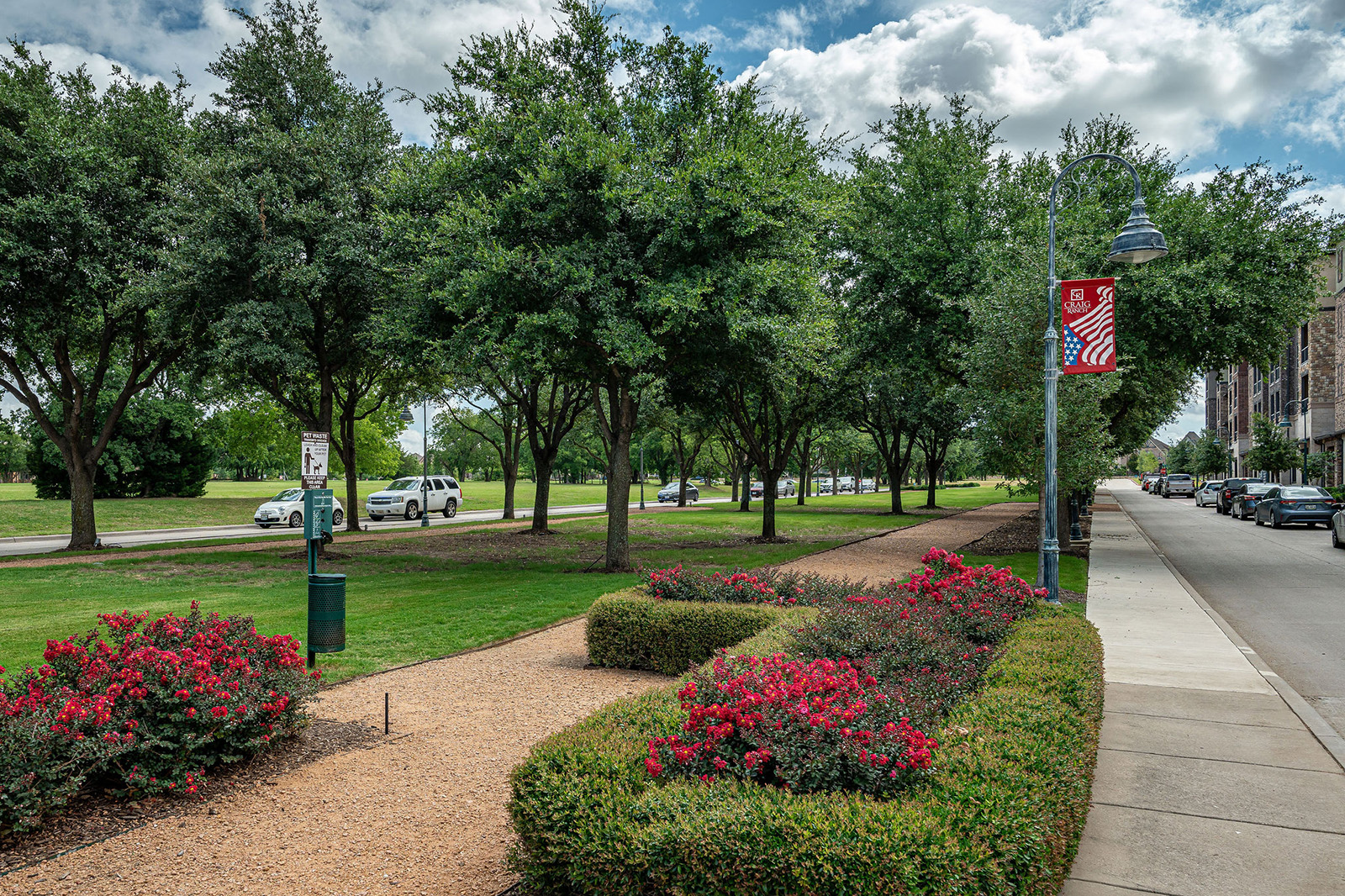 A landscaped sidewalk runs alongside a street lined with cars and trees, with red flowers in the foreground and a park area on the left.
