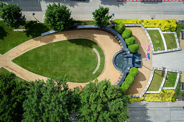 Aerial view of a landscaped outdoor memorial with a curved walkway, green lawn, central fountain, and an American flag near the entrance.