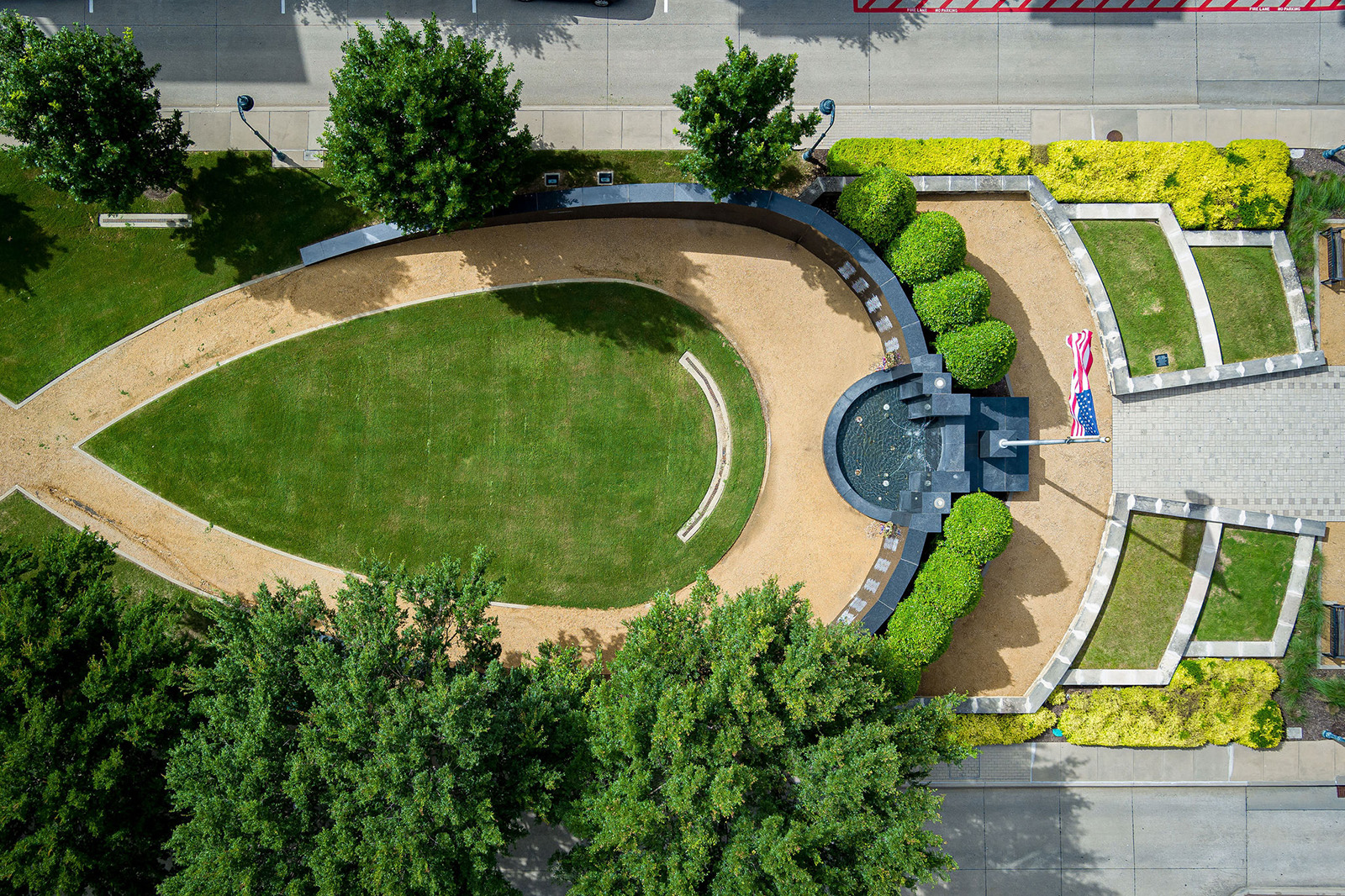 Aerial view of a landscaped memorial park with a curved pathway, central fountain, benches, green lawn, trees, and an American flag.