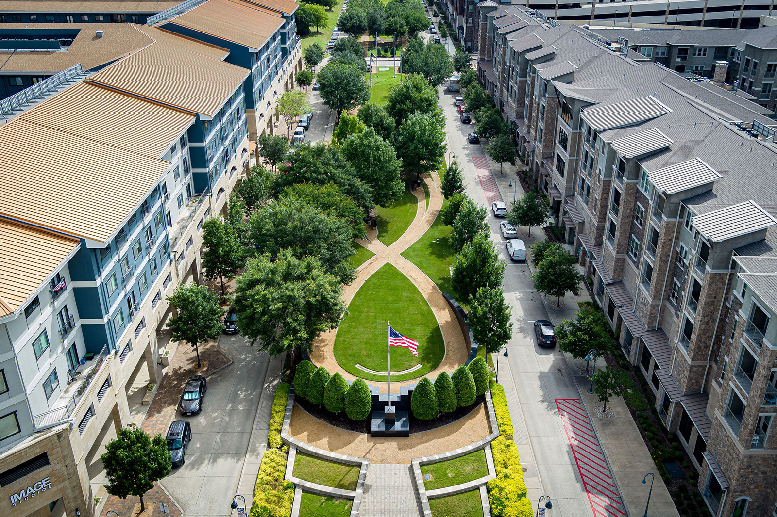 Aerial view of a landscaped courtyard with an American flag, surrounded by multi-story apartment buildings and parked cars on both sides.