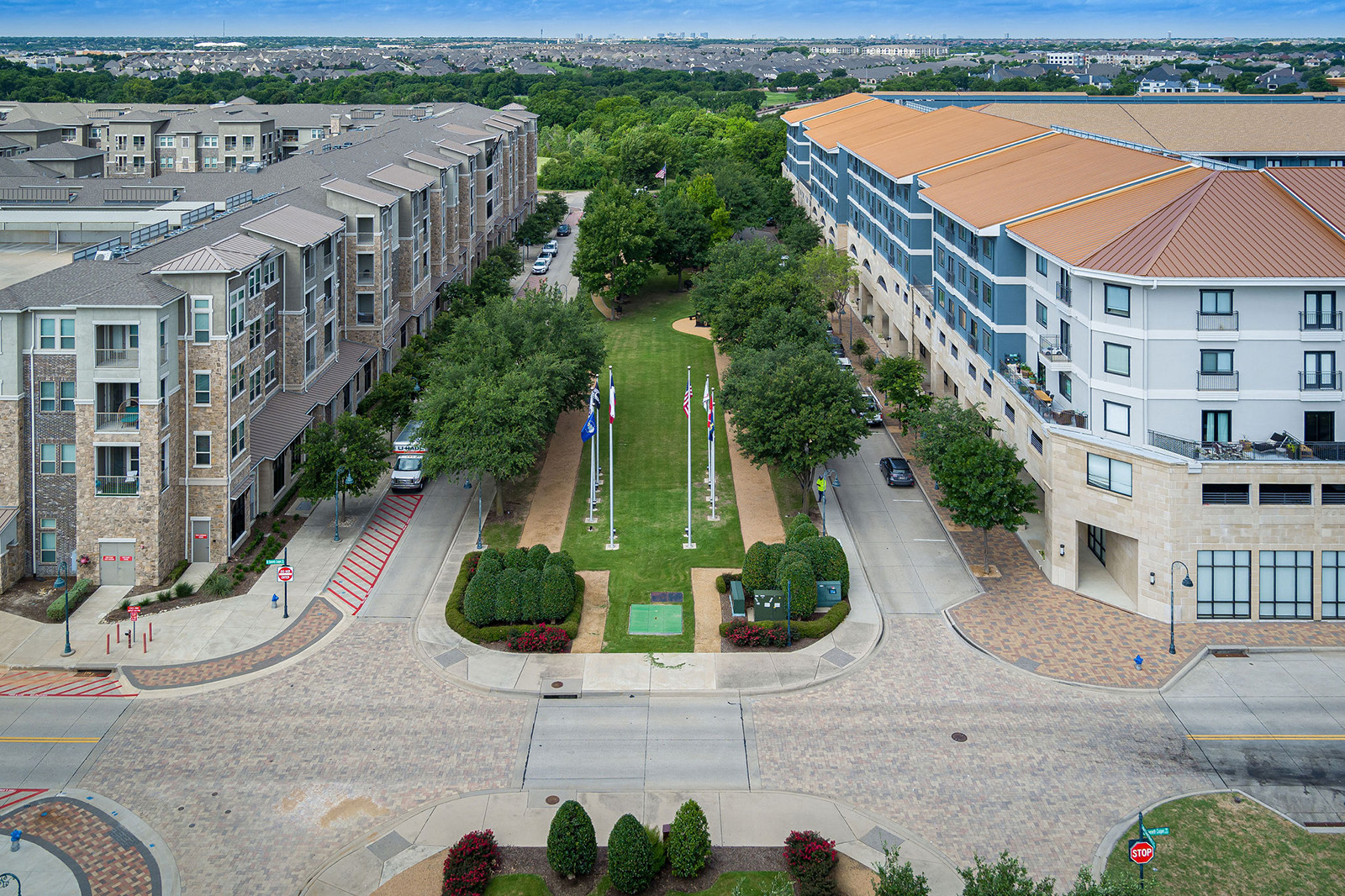 Aerial view of a landscaped plaza with flagpoles, surrounded by mid-rise apartment buildings and trees, with a cityscape visible in the distant background.