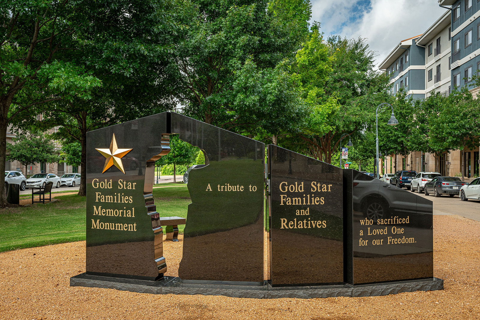 Gold Star Families Memorial Monument with gold lettering and a star, honoring families and relatives who lost loved ones in military service, set in a park area with trees and parked cars.