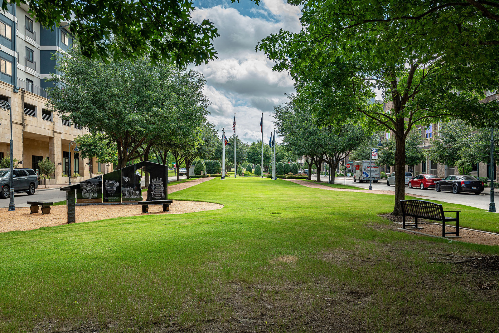 A grassy park area bordered by trees with flagpoles, a monument, benches, and buildings lining the street on both sides.